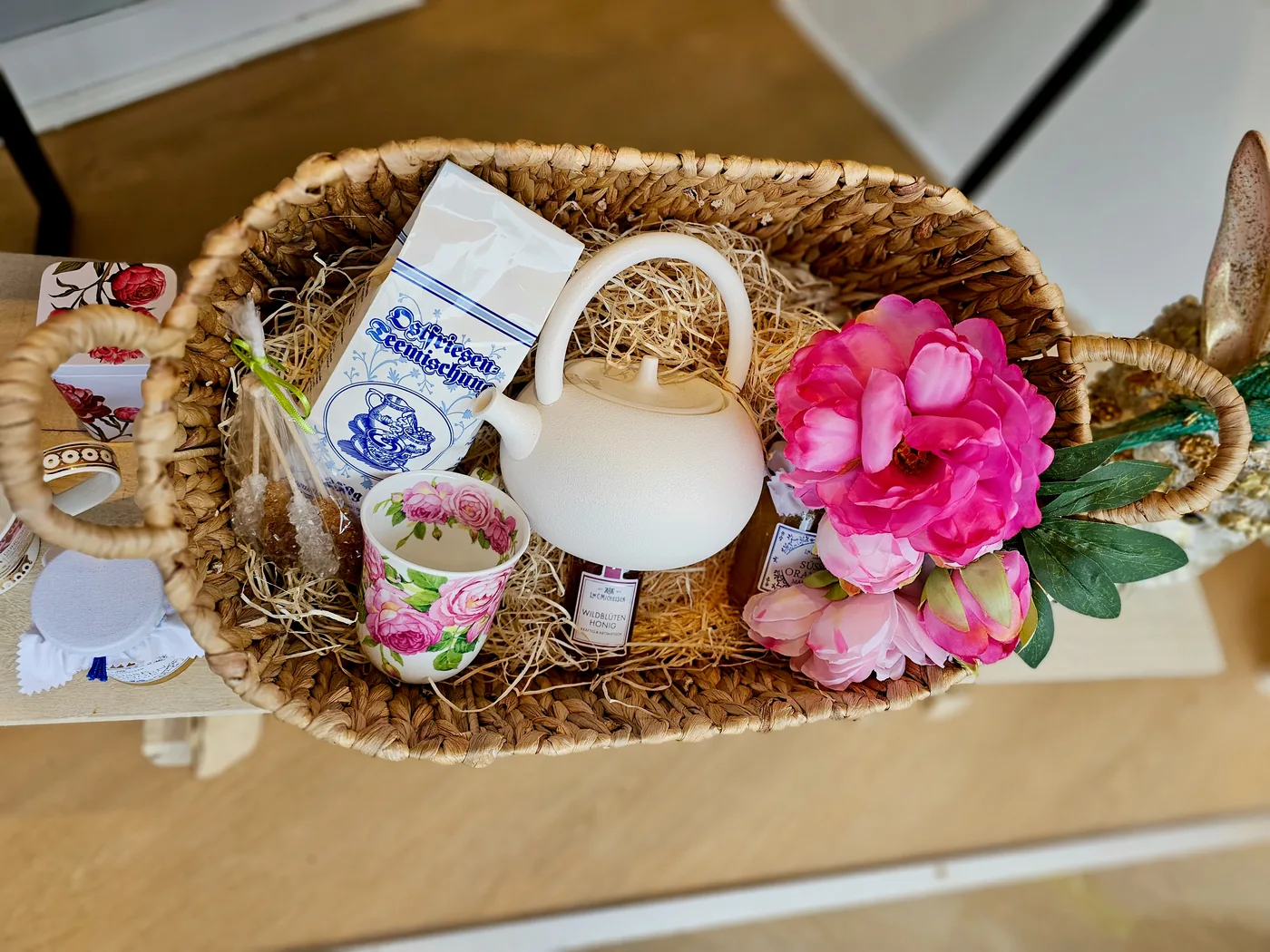 Una cesta de regalo trenzada con tetera blanca, taza de rosas, miel y té, decorada con flores rosas