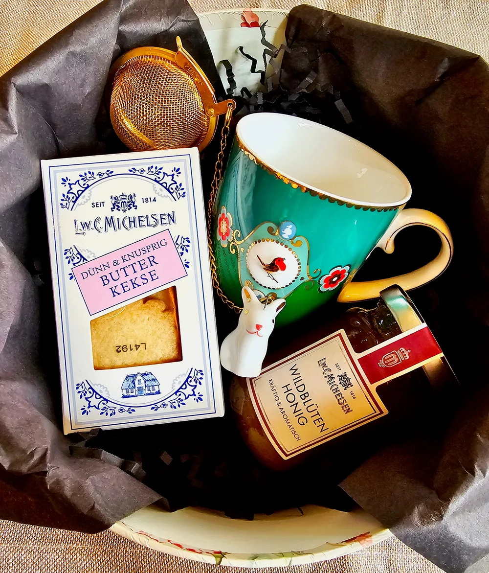 Una taza turquesa con un colador de té dorado, galletas de mantequilla y miel de flores silvestres sobre papel de seda negro