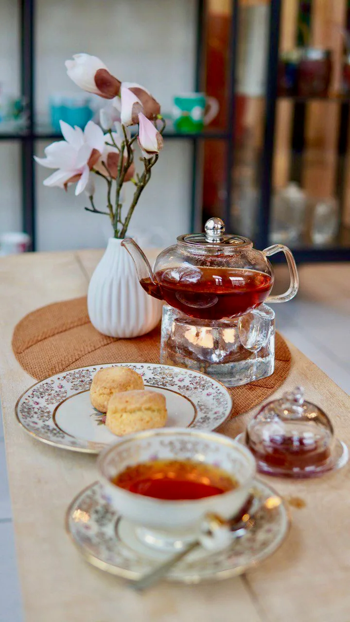 A teapot on a warmer, floral porcelain and scones beside magnolia blossoms