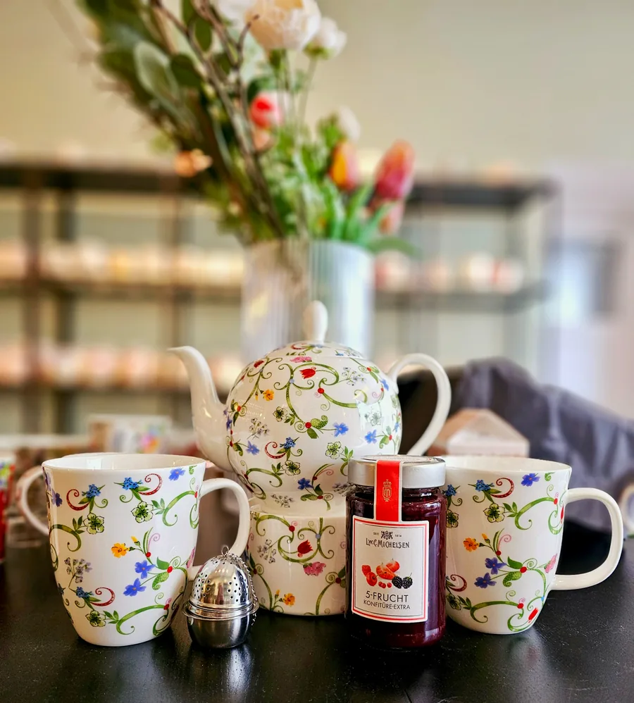 A white teapot and two cups with delicate floral decoration, a tea strainer and fruit jam