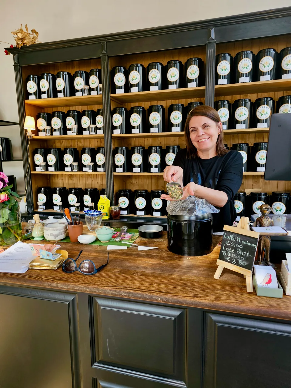 Maria Moreno holding a scoop of loose tea behind her shop counter, surrounded by tea tins