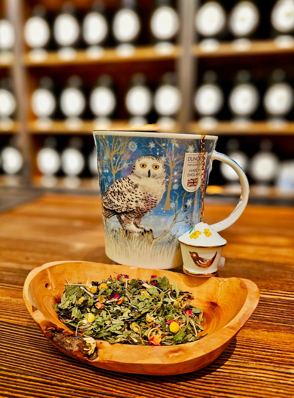 Una taza azul de porcelana Dunoon con un búho nival y un pequeño colador junto a un cuenco de madera con infusión de hierbas a granel