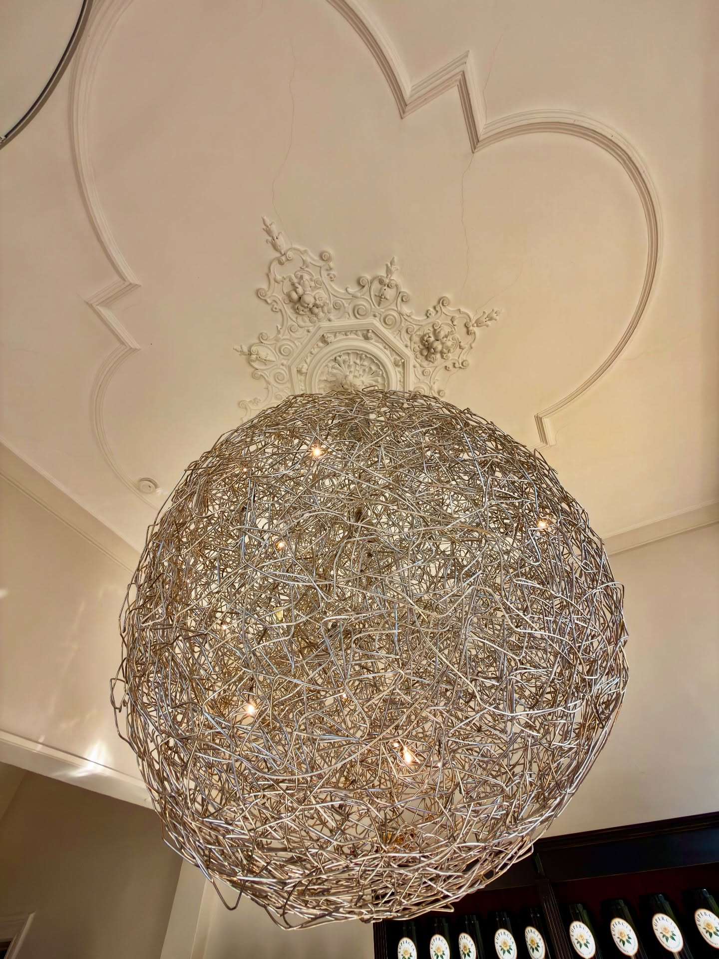 Skulpturaler Drahtkugel-Lüster vor der ornamentalen Stuckrosette der historischen Decke