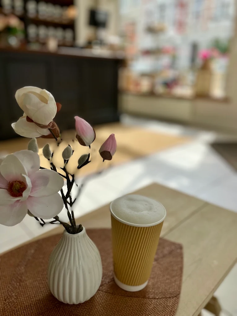 Magnolienzweig in geriffelter weißer Vase neben einem To-Go-Becher mit Milchschaum, sanft beleuchteter Tresen im Hintergrund